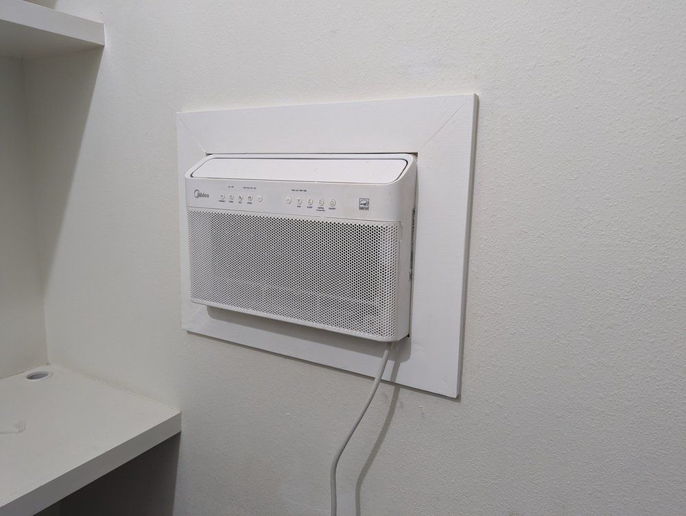 White air conditioner mounted in a wall, with a white frame. A cable hangs down.
