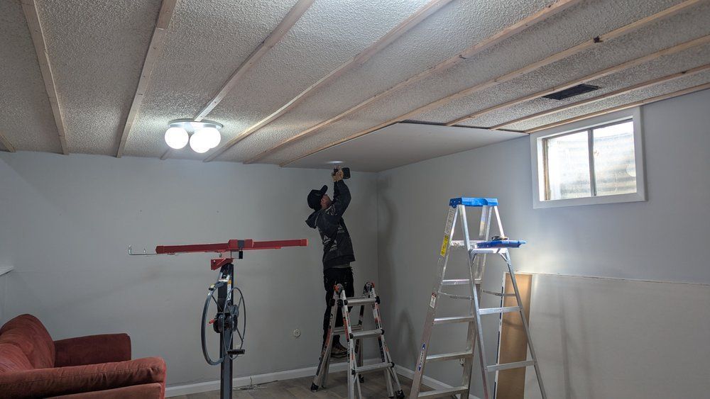 Person on ladder installs ceiling drywall in a basement. A lift and other tools are present.