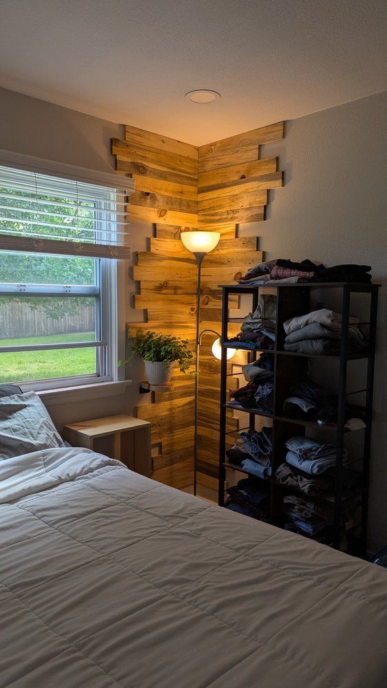 Bedroom corner with wooden accent wall, lamp, shelf with clothes, and a window.