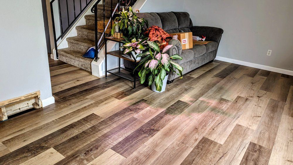 Living room with wood-look flooring, gray couch, stairs, plants, and a black metal shelf.