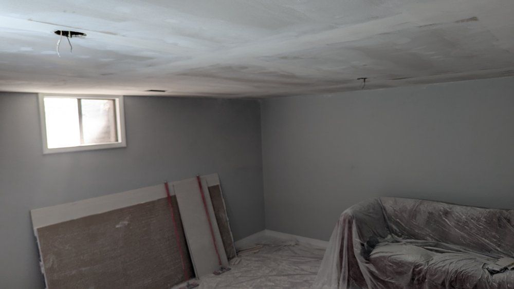 Room under construction, with gray walls, unfinished ceiling, window, and covered sofa. Drywall sheets are propped up.