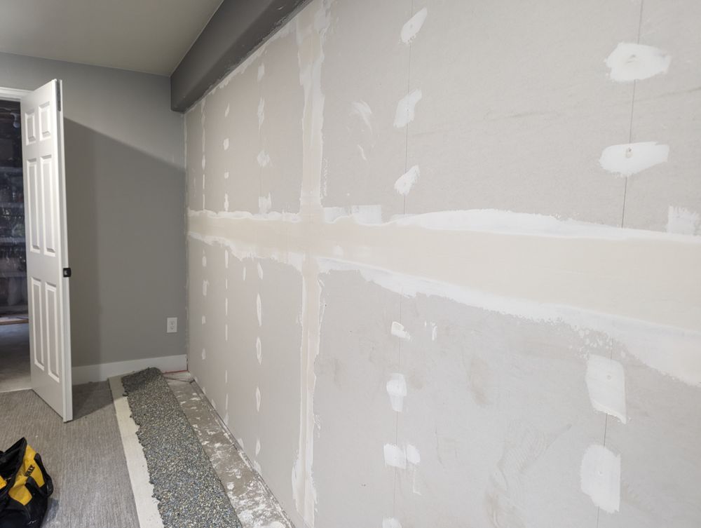 Drywall installation in progress. Interior wall with taped seams, patches of spackle, and door frame.