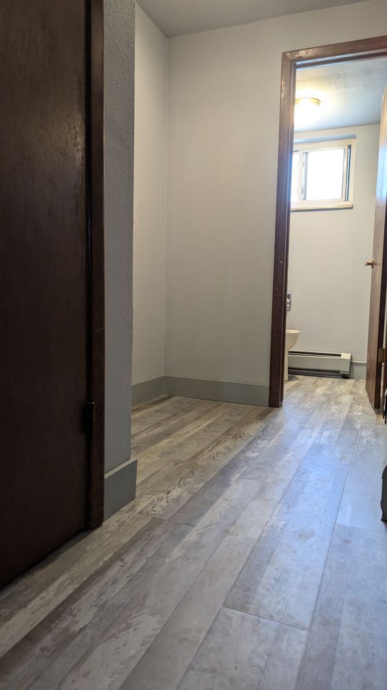 Narrow hallway with wood-look floor, doorways on either side, and a bathroom at the end with a window.