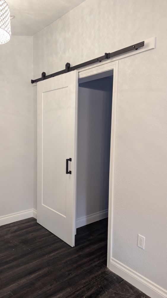 White barn door, black hardware, partially open revealing a doorway, on dark wood flooring.