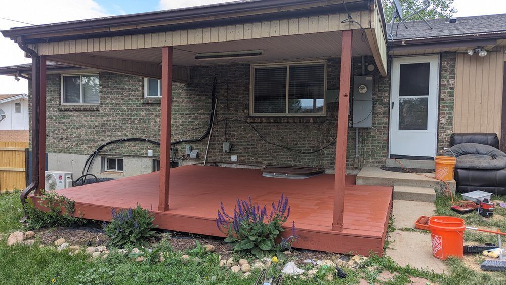 Red deck with overhead cover attached to a brick house. Yard with tools, bushes.