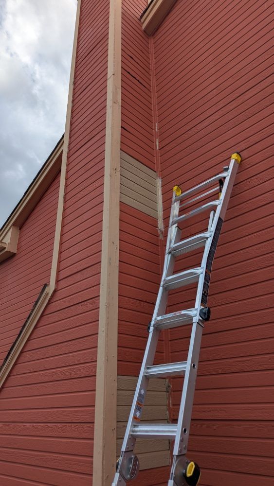 Ladder leaning against a red building with light wood trim and patch. Overcast sky.