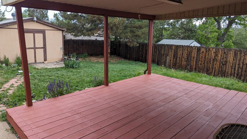 Red wooden deck under a covered porch overlooking a backyard with a shed and wooden fence.