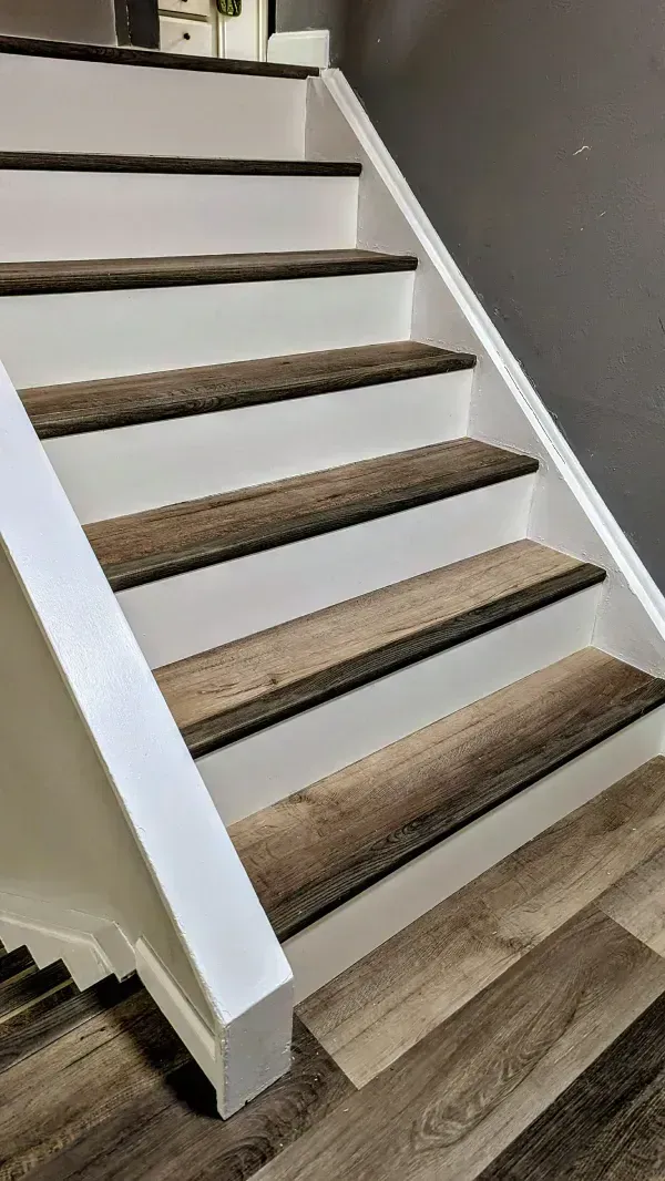 Staircase with white risers and wood-look treads. The stairs lead upwards against a grey wall and white railing.