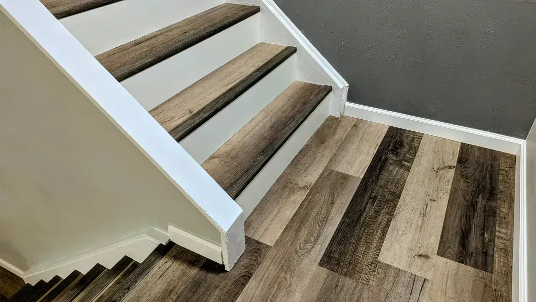 Staircase with white risers and wood-look treads. The stairs lead to a kitchen with white cabinets. Gray wall.