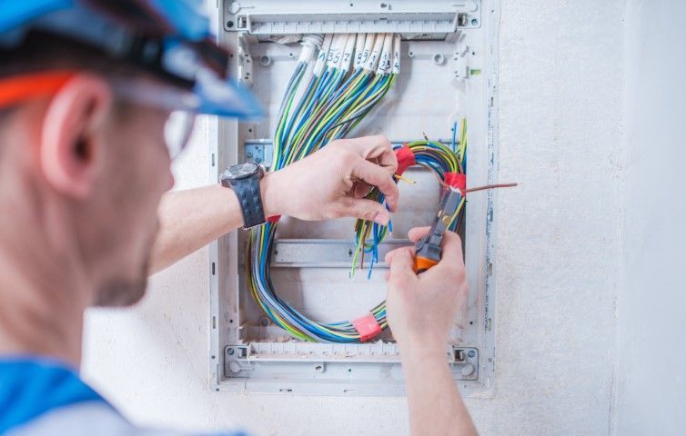 Electrician working on electrical wiring in a panel, wearing safety glasses and a hard hat.