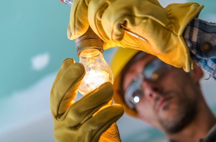 Electrician in a yellow hard hat and gloves screwing in a light bulb.
