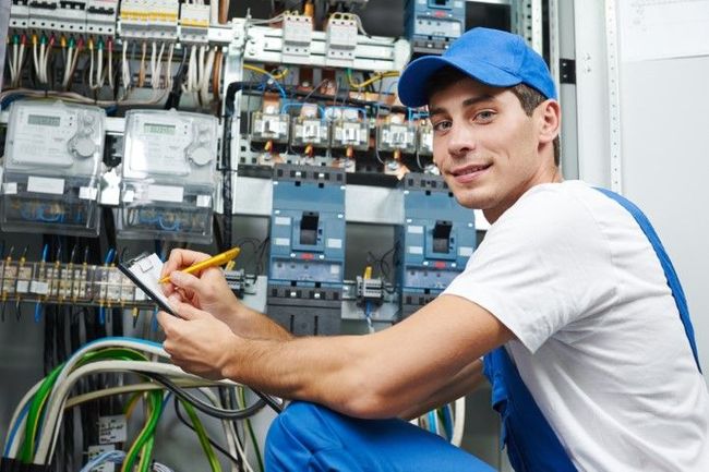 Electrician in blue uniform writing on clipboard, inspecting electrical panel.