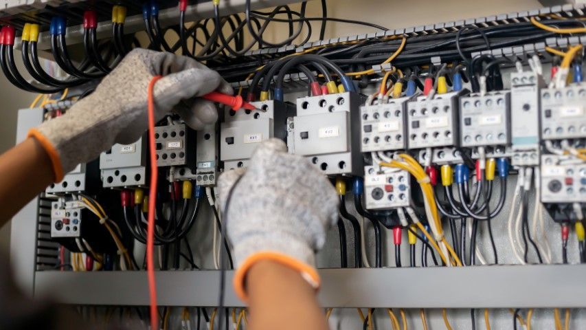 Person in gloves testing electrical panel wiring with a multimeter.