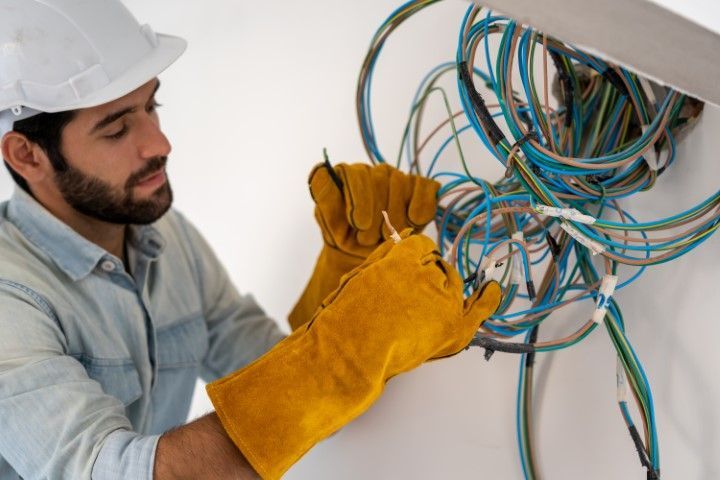Electrician working with wires, wearing a hard hat and gloves, on a white wall.