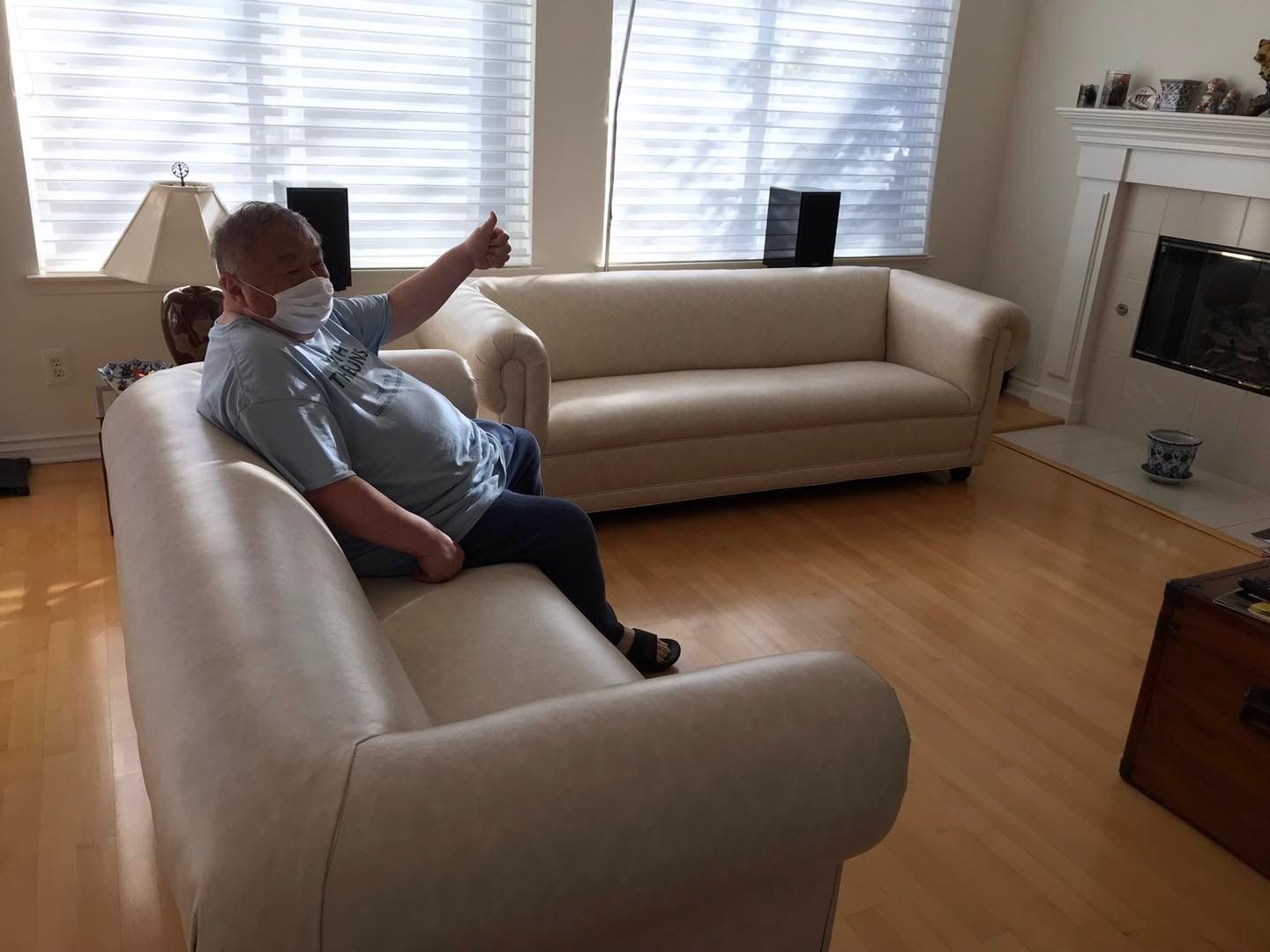 A man wearing a mask is sitting on a white couch in a living room.