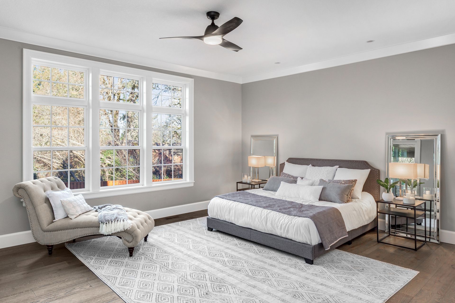 Chambre avec grandes fenêtres, murs gris, lit, chaise longue et tapis.