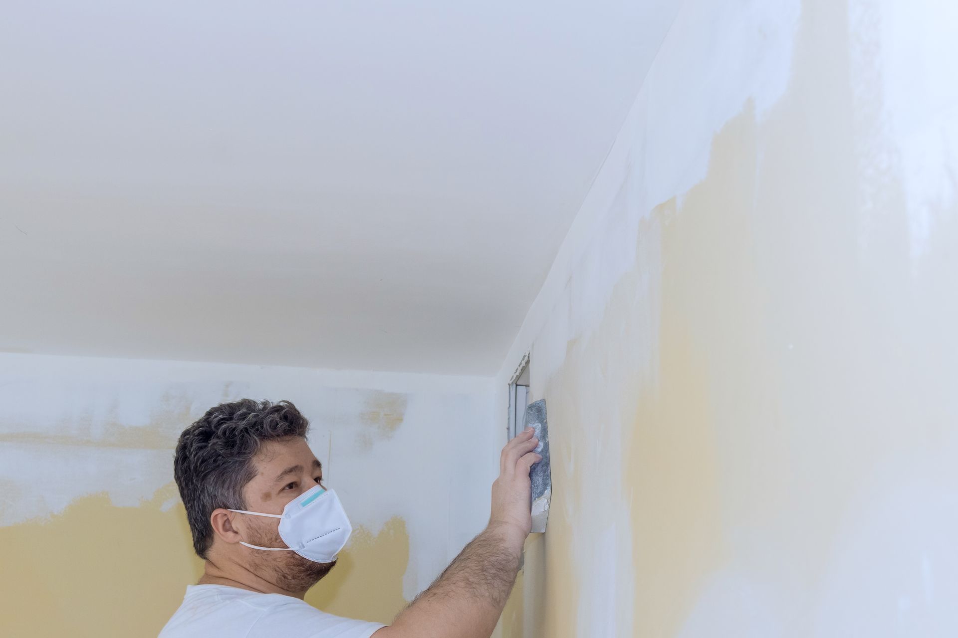 Un homme portant un masque utilise une truelle pour appliquer du composé pour cloisons sèches sur un mur d'une pièce.