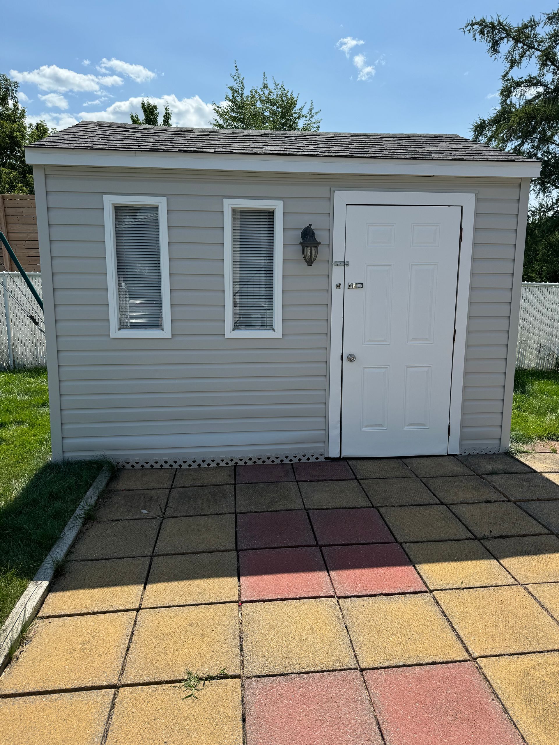 Petit cabanon gris clair avec porte blanche et deux fenêtres, sur un patio carrelé coloré.