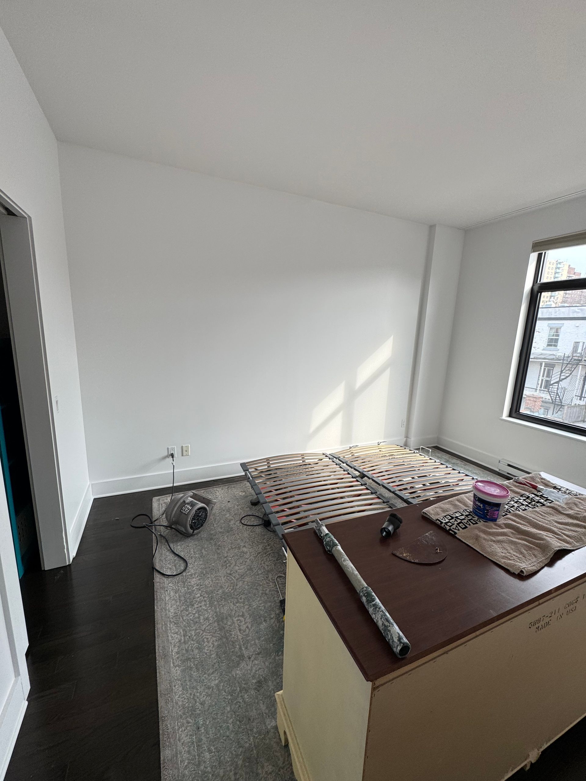 Pièce vide avec murs blancs et sol sombre, tapis et petite table au premier plan, fenêtre à droite.