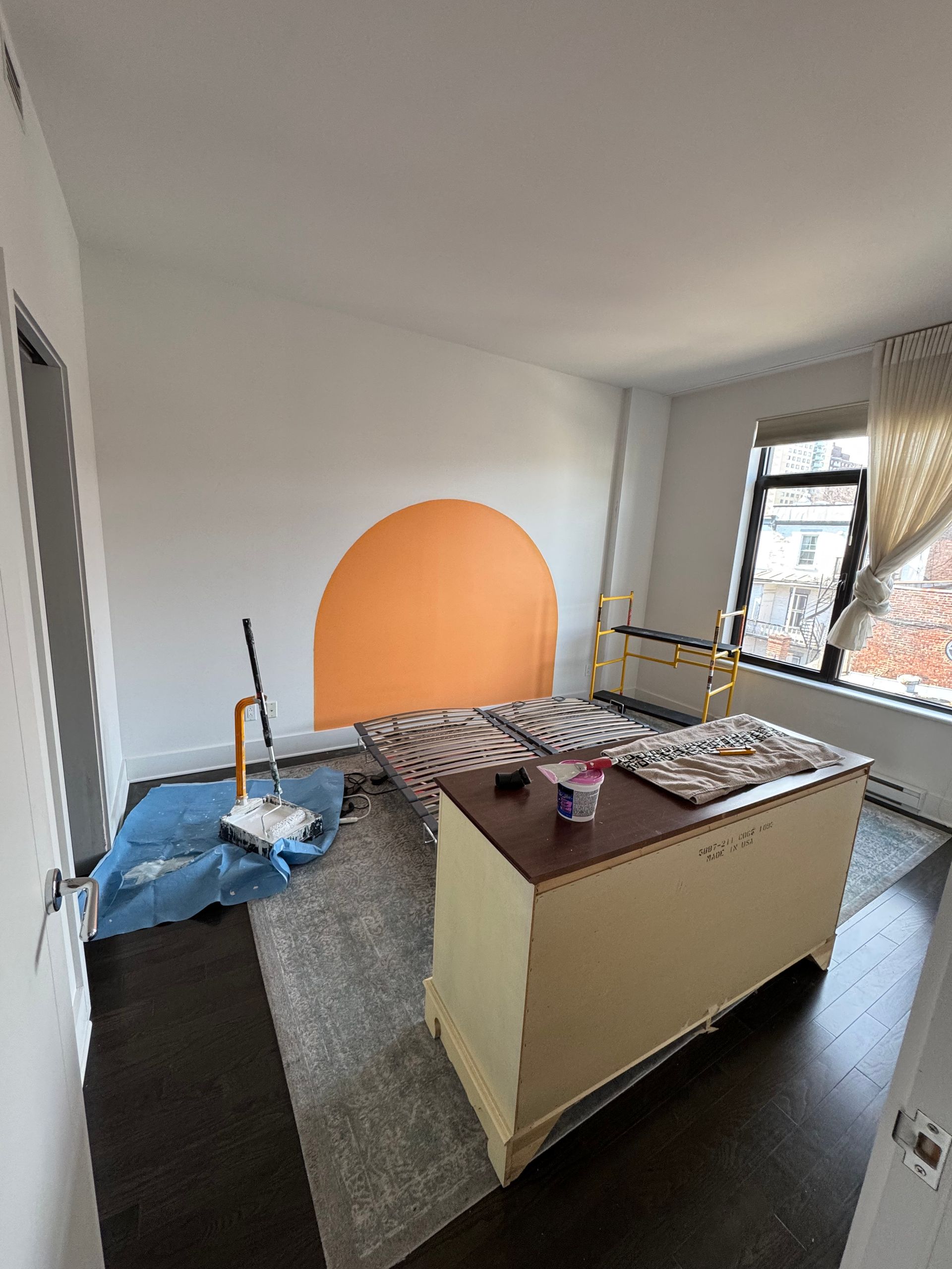 Une chambre avec un demi-cercle orange peint sur le mur, un lit, un coffre et des fournitures de peinture.