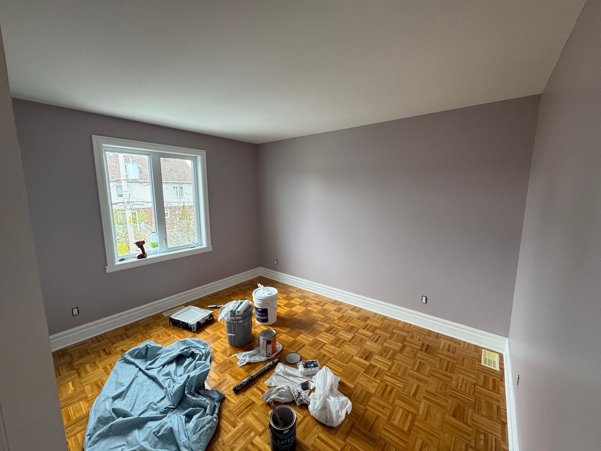 Pièce vide, peinte en violet clair, avec parquet, fenêtre et fournitures de peinture éparpillées.