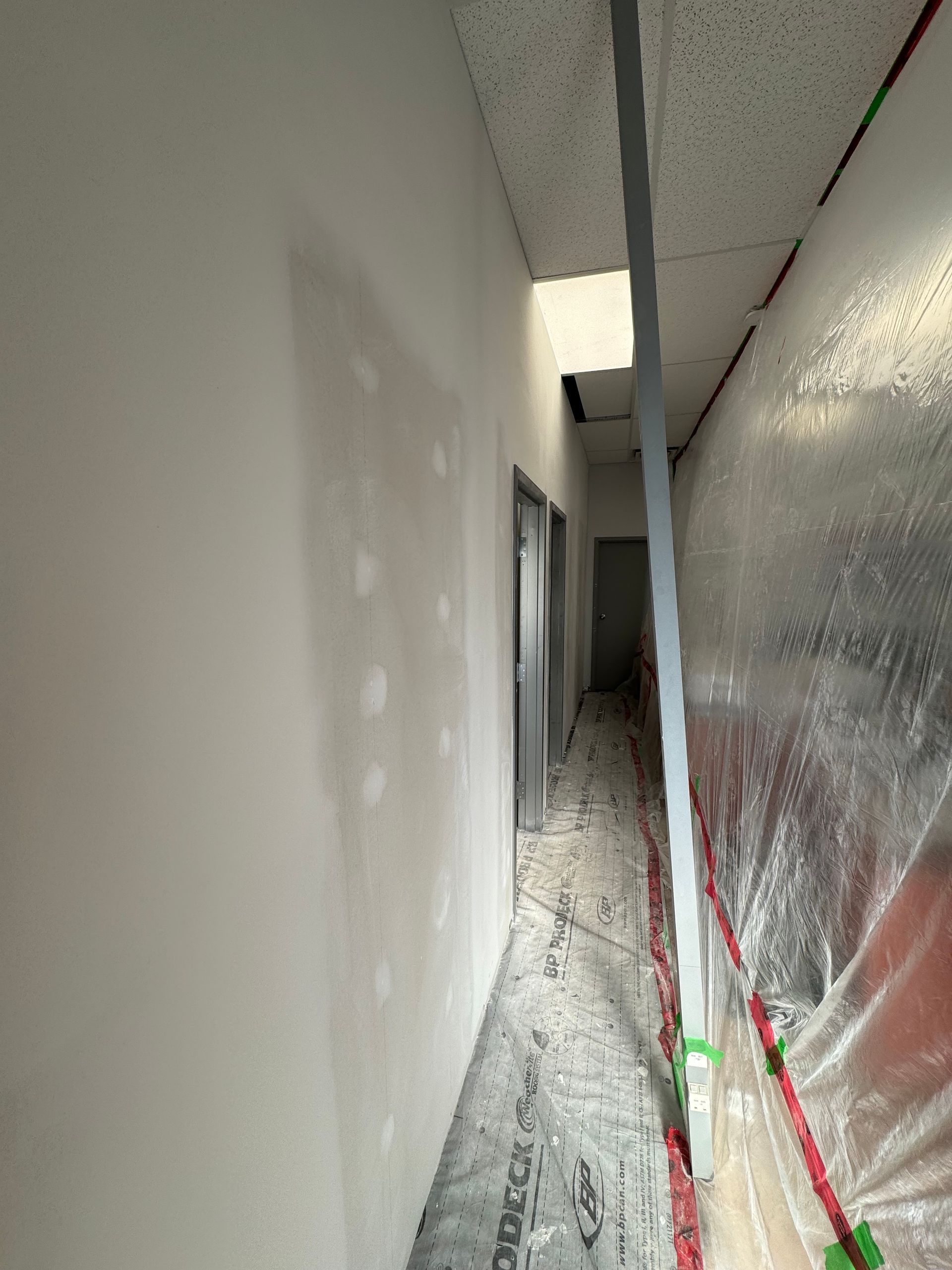 Couloir étroit et inachevé avec des murs blancs, une porte grise au bout et une bâche de protection à droite.