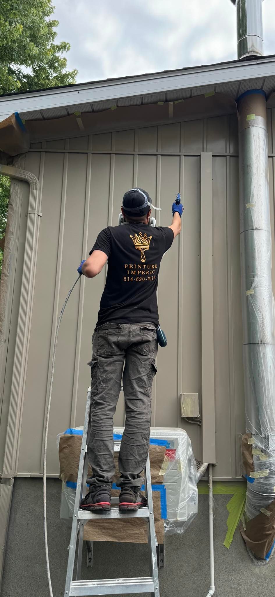 Une personne sur une échelle pulvérise du produit sur le côté d'un bâtiment. Elle porte un chapeau, un masque, des gants et un pantalon camouflage.