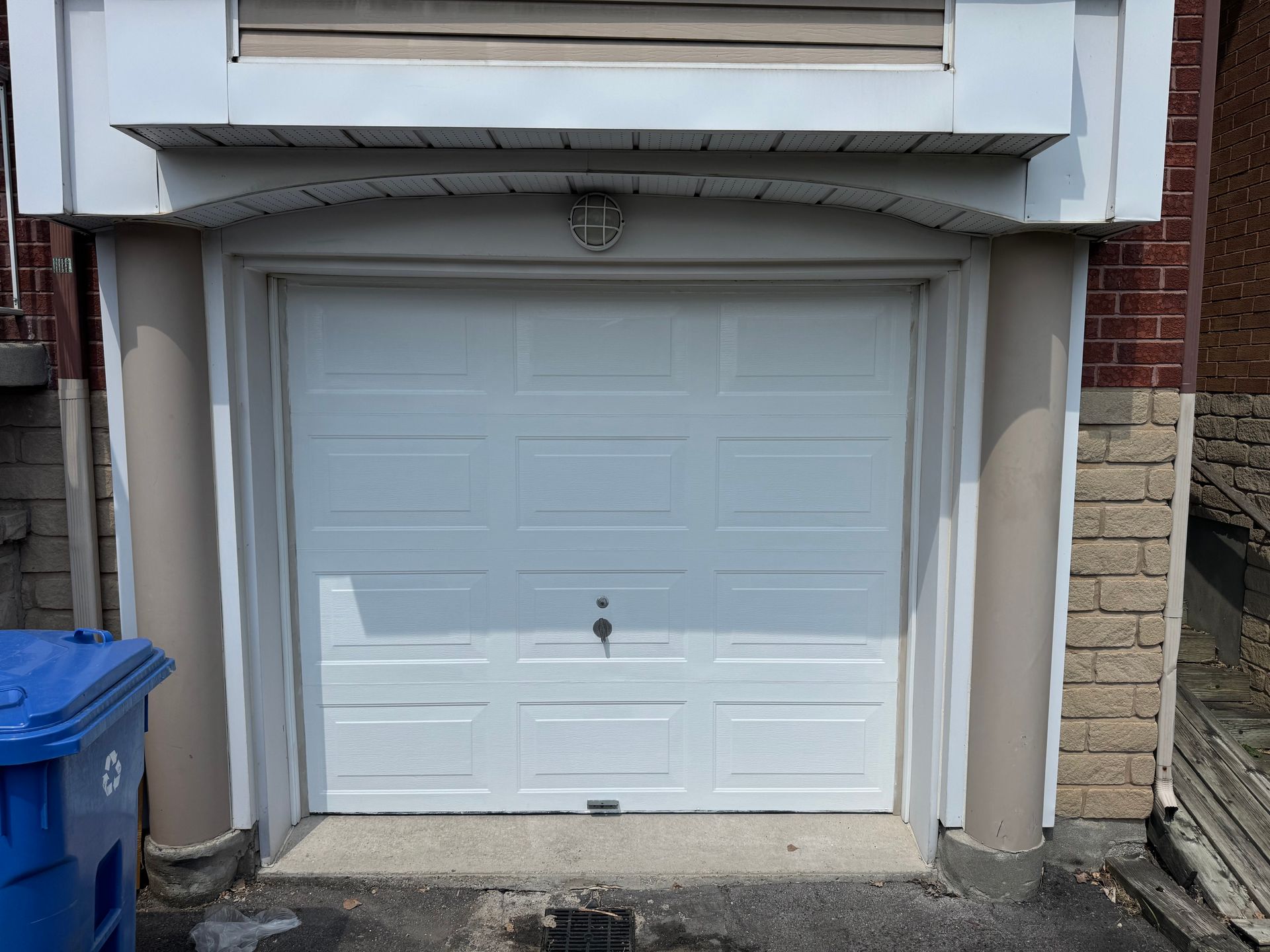 Porte de garage blanche avec un bac de recyclage bleu à gauche.