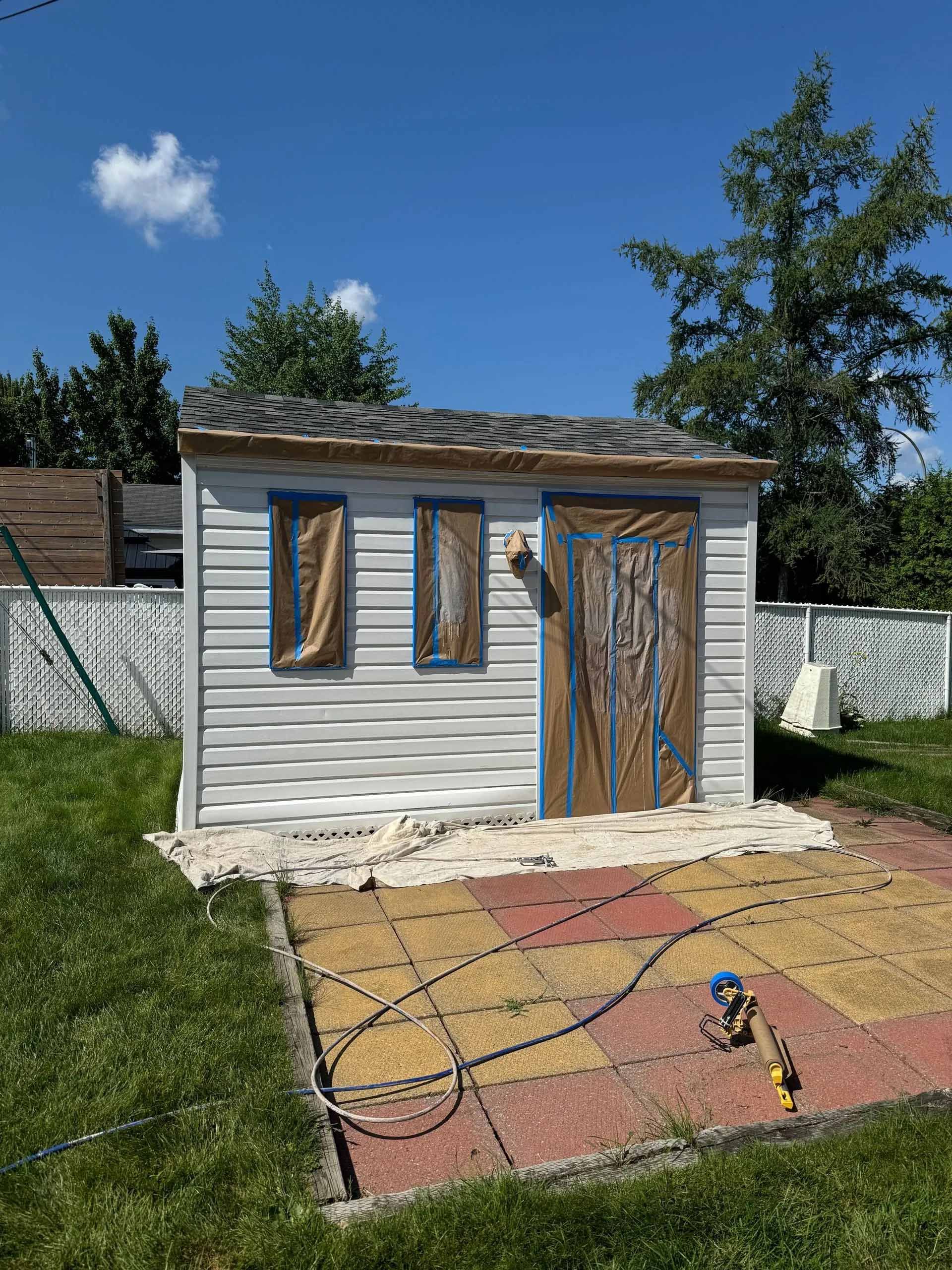 Un petit hangar blanc en cours de préparation pour la peinture avec du ruban adhésif bleu et du papier brun ; extérieur ensoleillé.