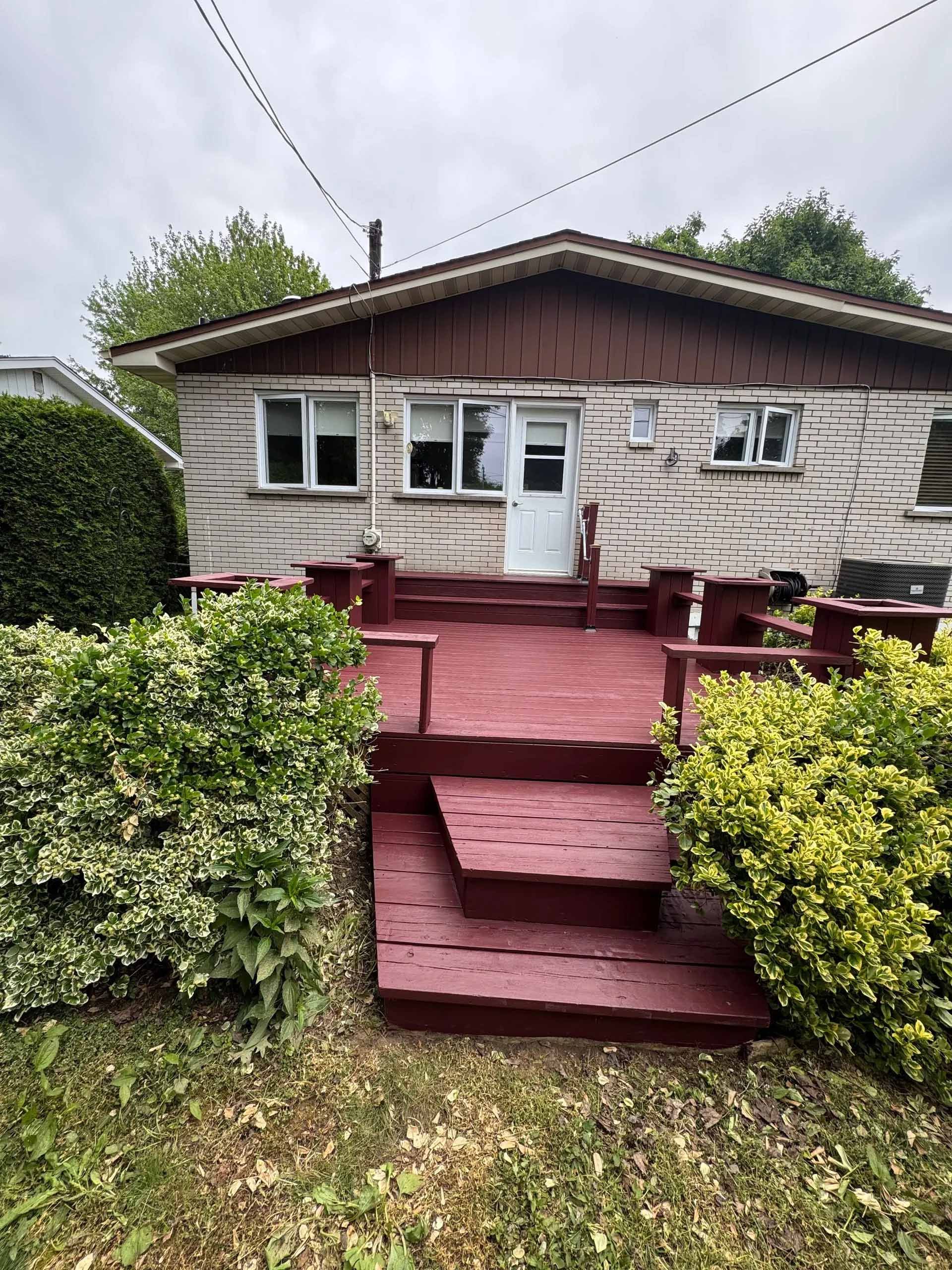 Maison avec revêtement en briques, terrasse rouge et buissons. Ciel couvert.