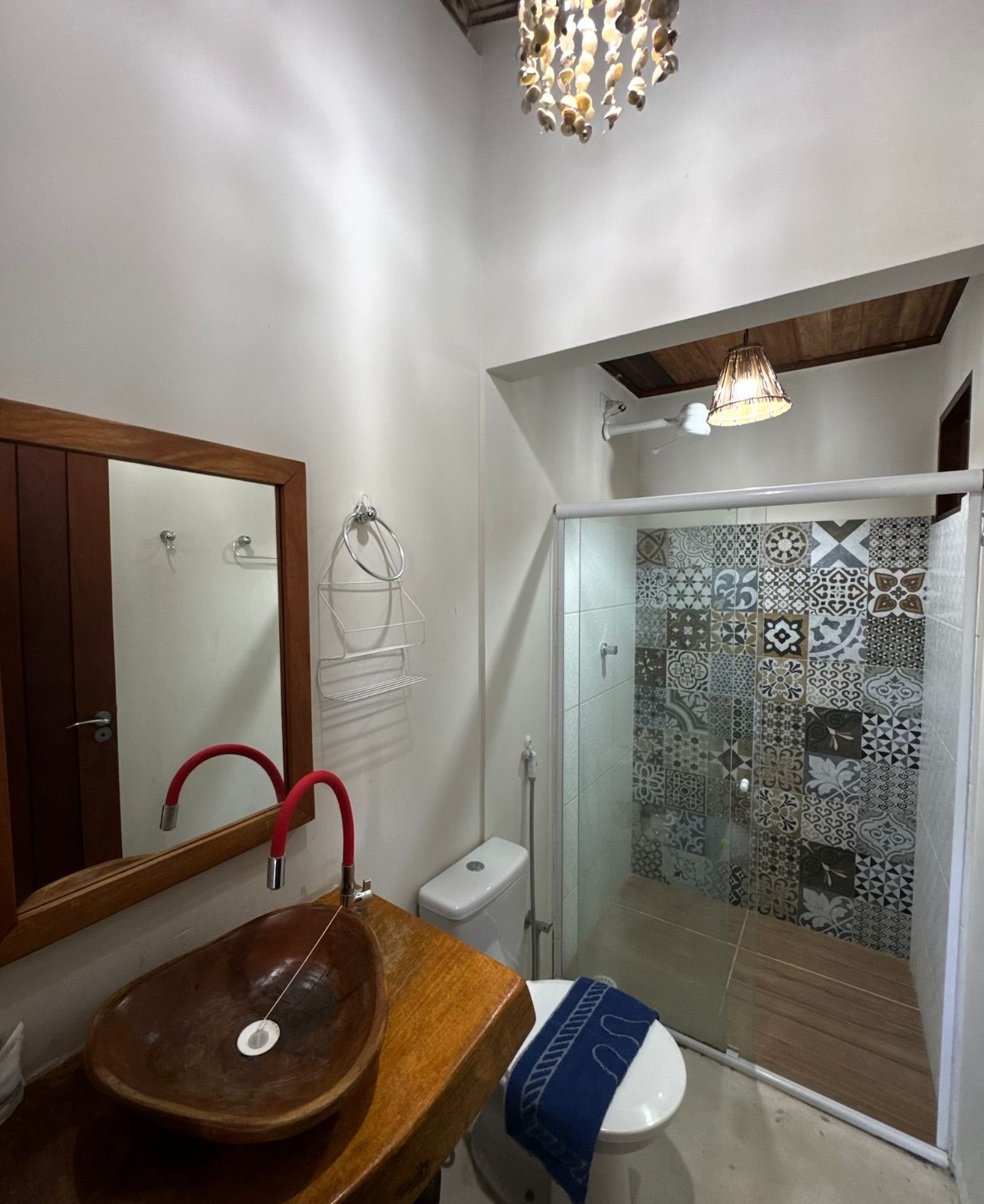 Um banheiro com uma bancada de madeira, uma pia de pedra com torneira vermelha, um vaso sanitário e um box de vidro com paredes revestidas de azulejos.