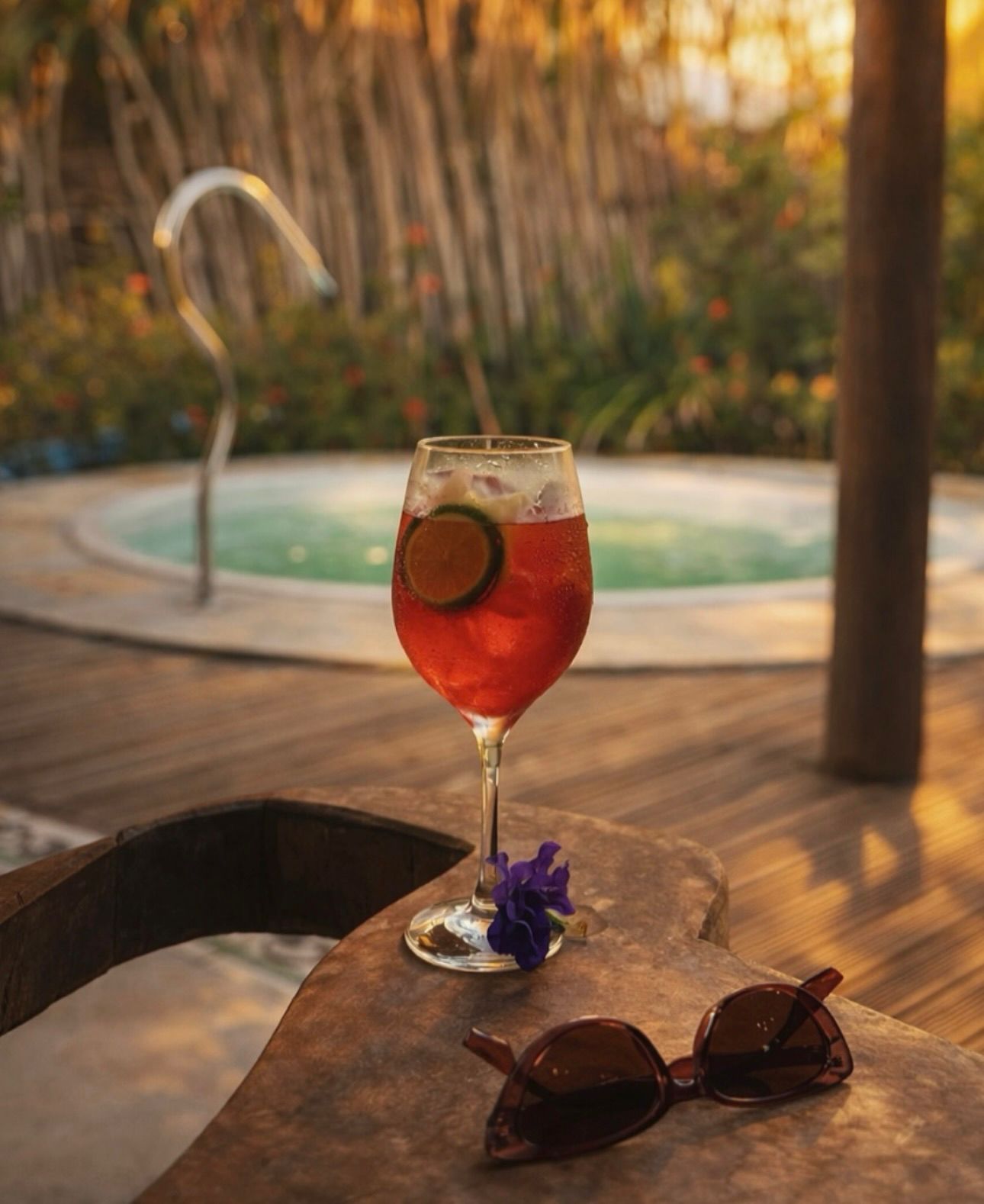 Um coquetel vermelho com uma rodela de limão repousa sobre uma mesa de pedra ao lado de óculos de sol, com uma piscina externa desfocada ao fundo.
