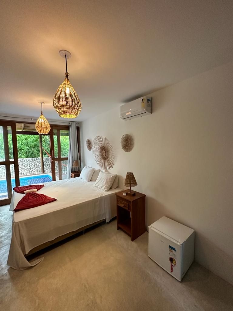 Um quarto de hotel com cama, mesa de cabeceira de madeira, frigobar branco, ar-condicionado de parede e luminárias pendentes.