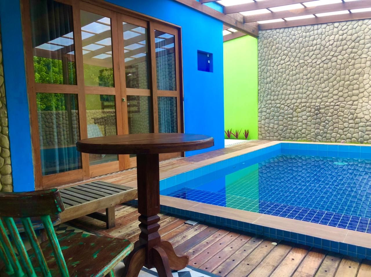 Uma mesa e uma cadeira de madeira em um deck ao lado de uma piscina azul vibrante, com paredes azuis e verdes e uma parede de pedra.