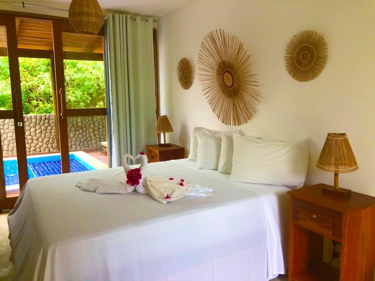 Um quarto de hotel com cama queen-size, decorado com cisnes feitos de toalhas, mesas de cabeceira, parede revestida de vime e vista para a piscina através de uma janela de vidro.