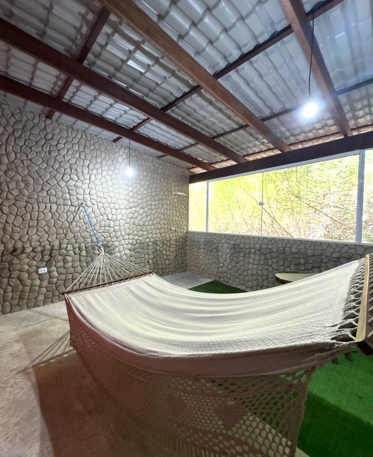 Uma rede está pendurada dentro de um pátio coberto ao ar livre, com paredes de pedra texturizada e piso de grama verde.