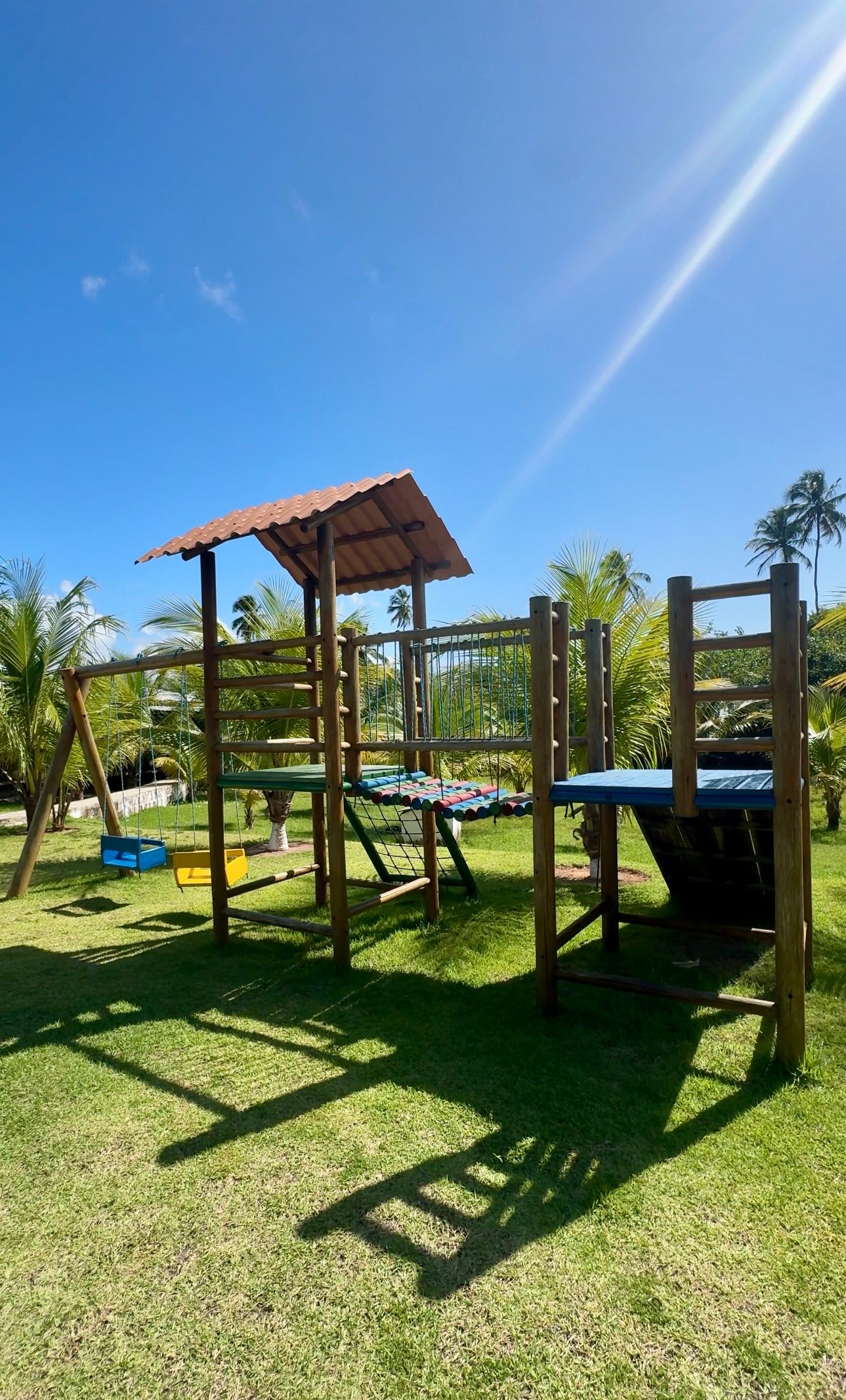 Uma estrutura de madeira para parque infantil, com cobertura, plataformas verdes e balanços, ergue-se num gramado sob um céu azul brilhante.