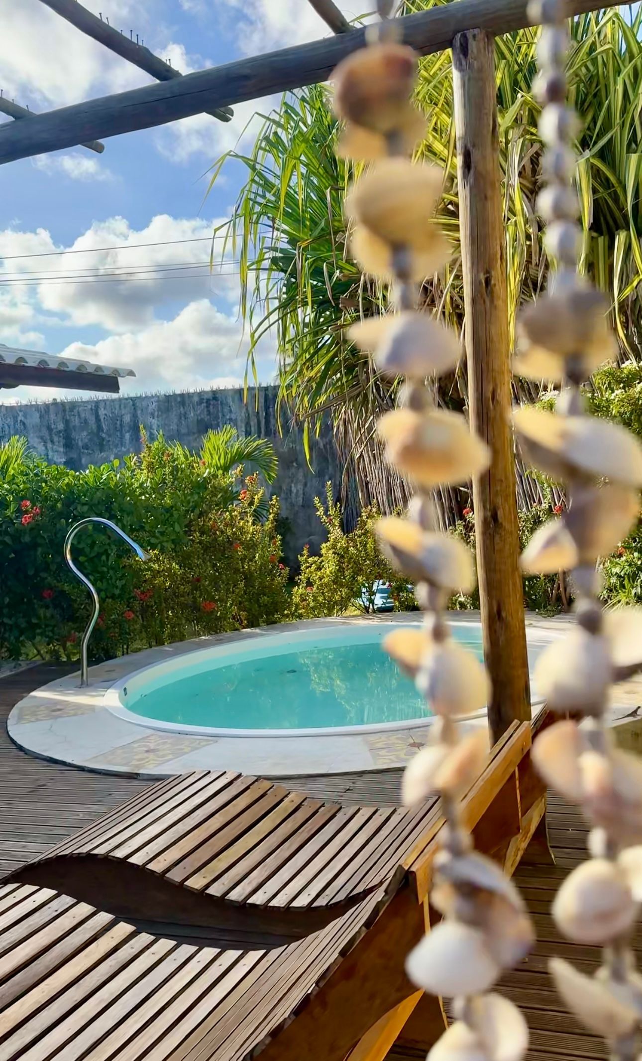 Uma piscina circular rodeada de vegetação e espreguiçadeiras de madeira, visível através de um fio de conchas suspensas.