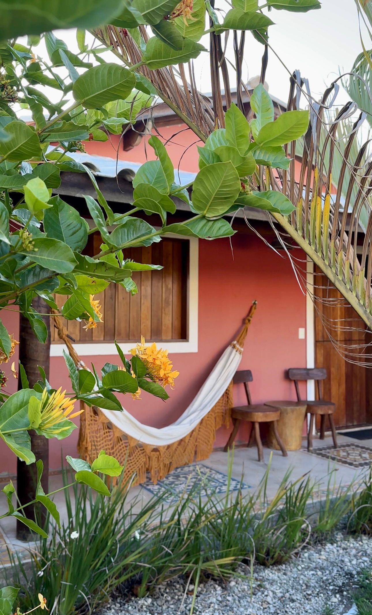 Uma rede branca está pendurada na varanda de um prédio cor salmão, acompanhada por duas cadeiras de madeira perto de uma árvore verde.