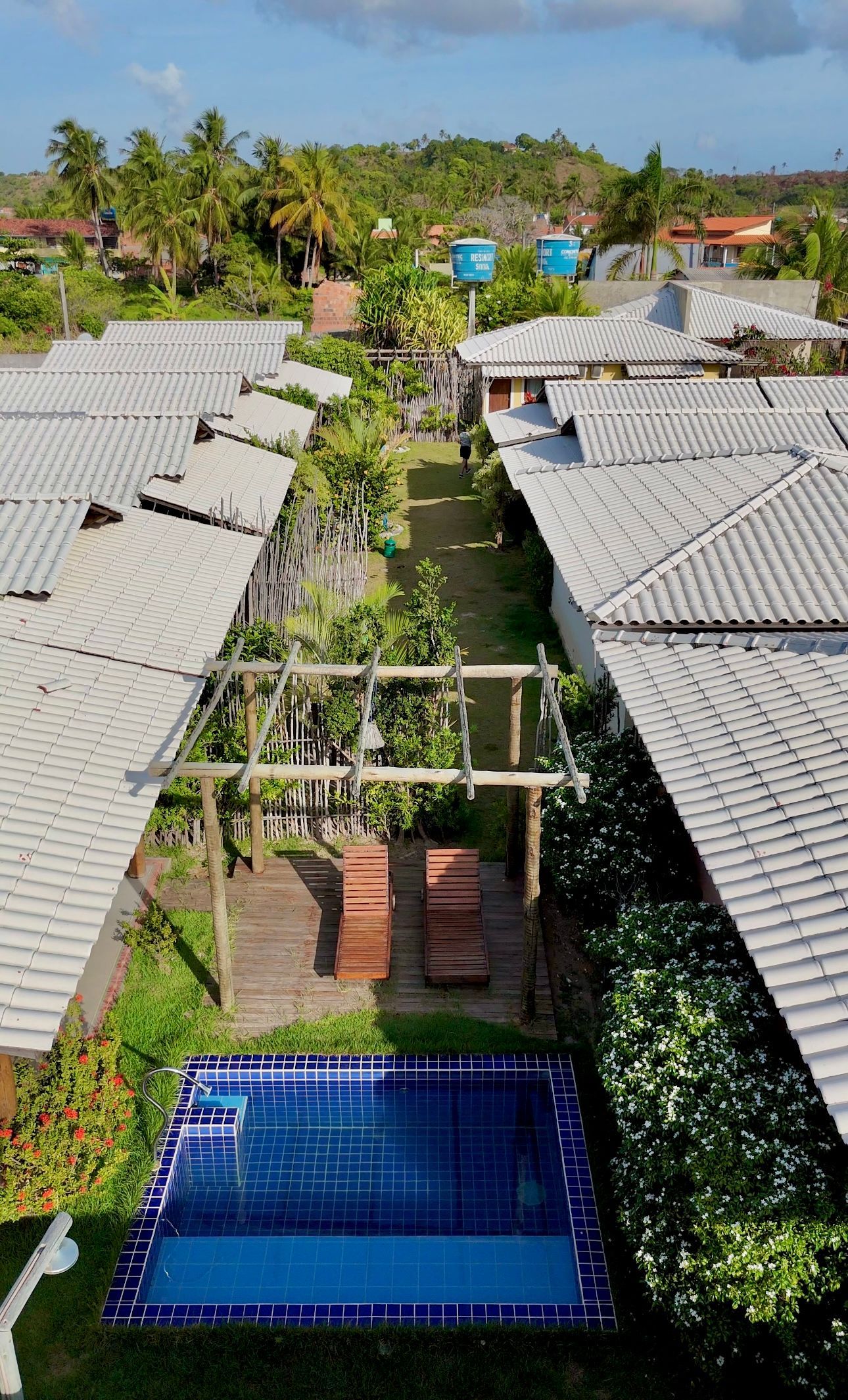 Um pátio com uma piscina revestida de azulejos azuis, duas espreguiçadeiras de madeira sob um pergolado e fileiras de casas com telhados cinzentos.