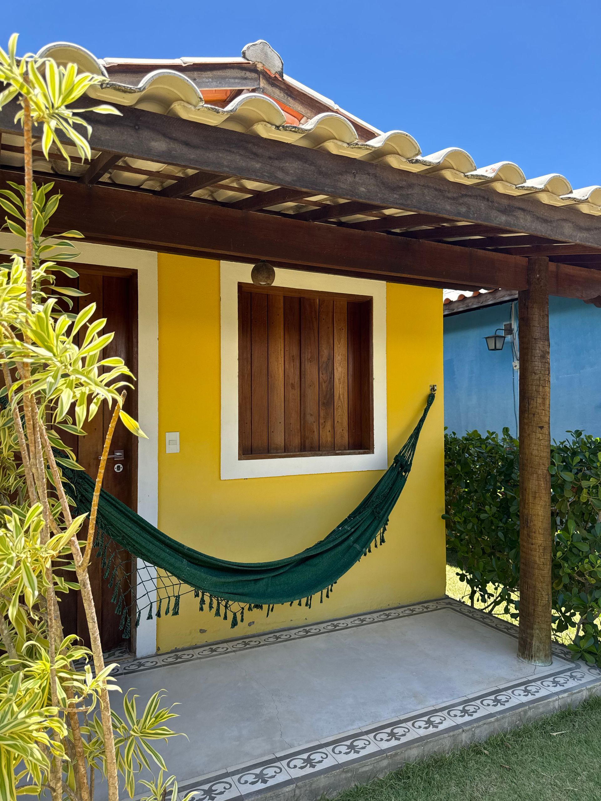 Uma casinha amarela com uma rede verde na varanda, com uma janela de madeira e um telhado de telhas sob um céu azul claro.