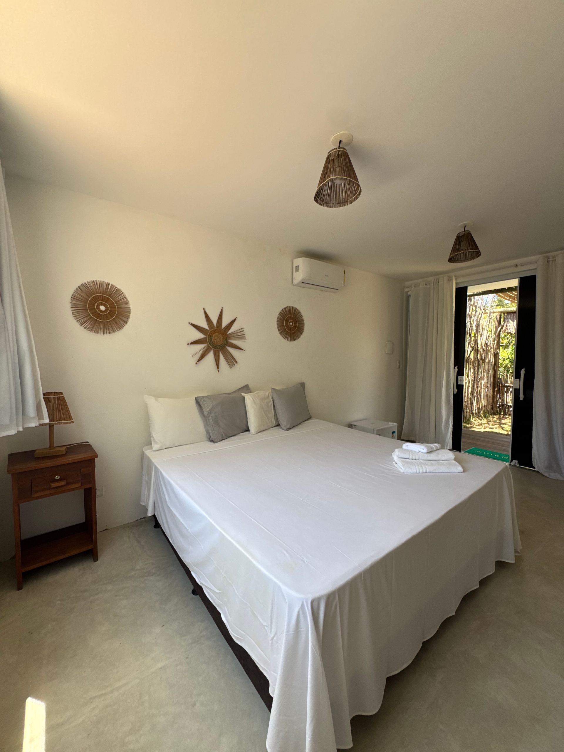 Um quarto de hotel minimalista com cama branca, paredes bege com decoração em formato de sol, dois lustres pendentes de vime e uma porta de vidro.