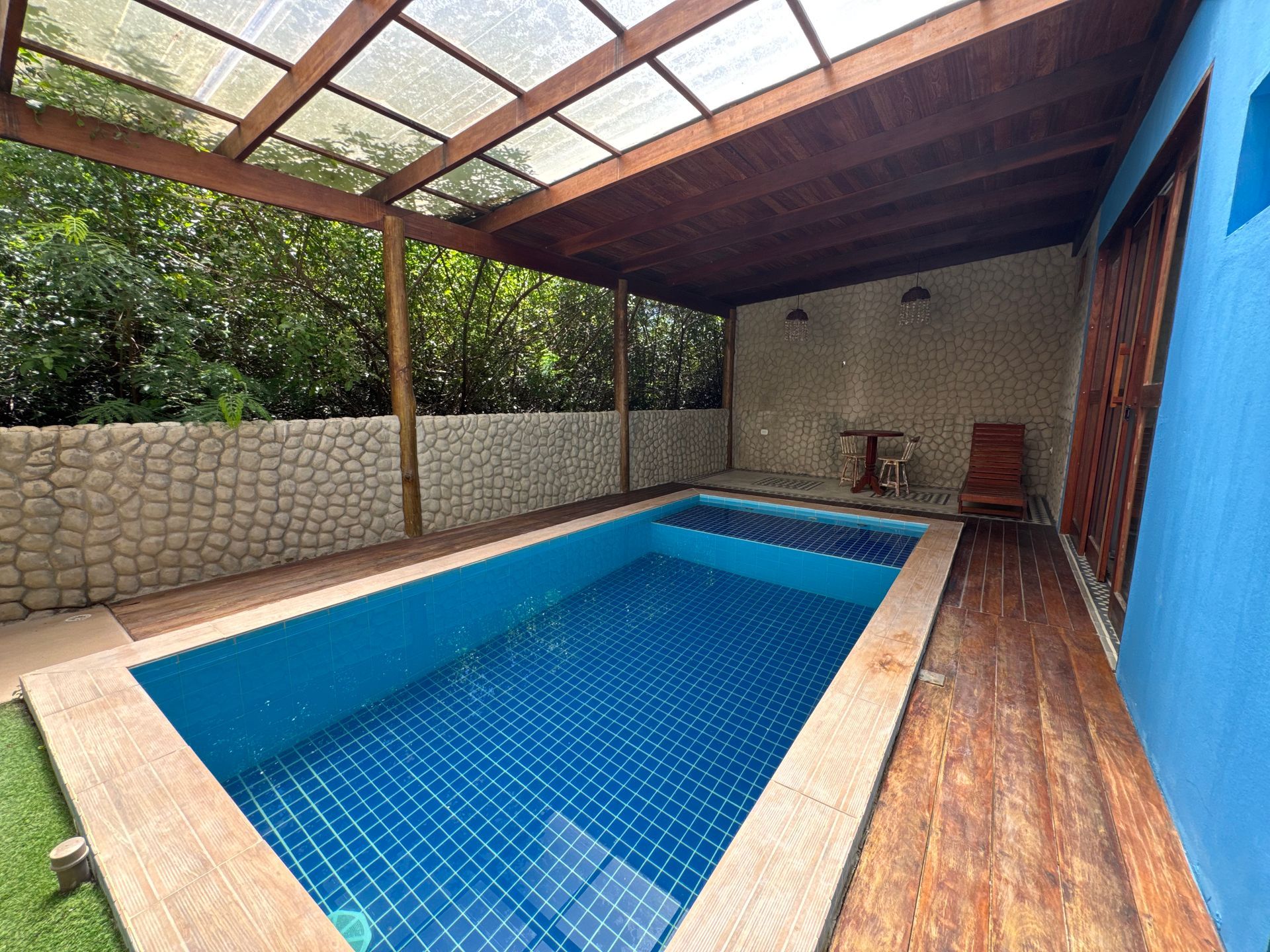 Uma piscina retangular com azulejos azuis fica sob uma pérgola de madeira em um deck de madeira ao lado de uma parede azul.