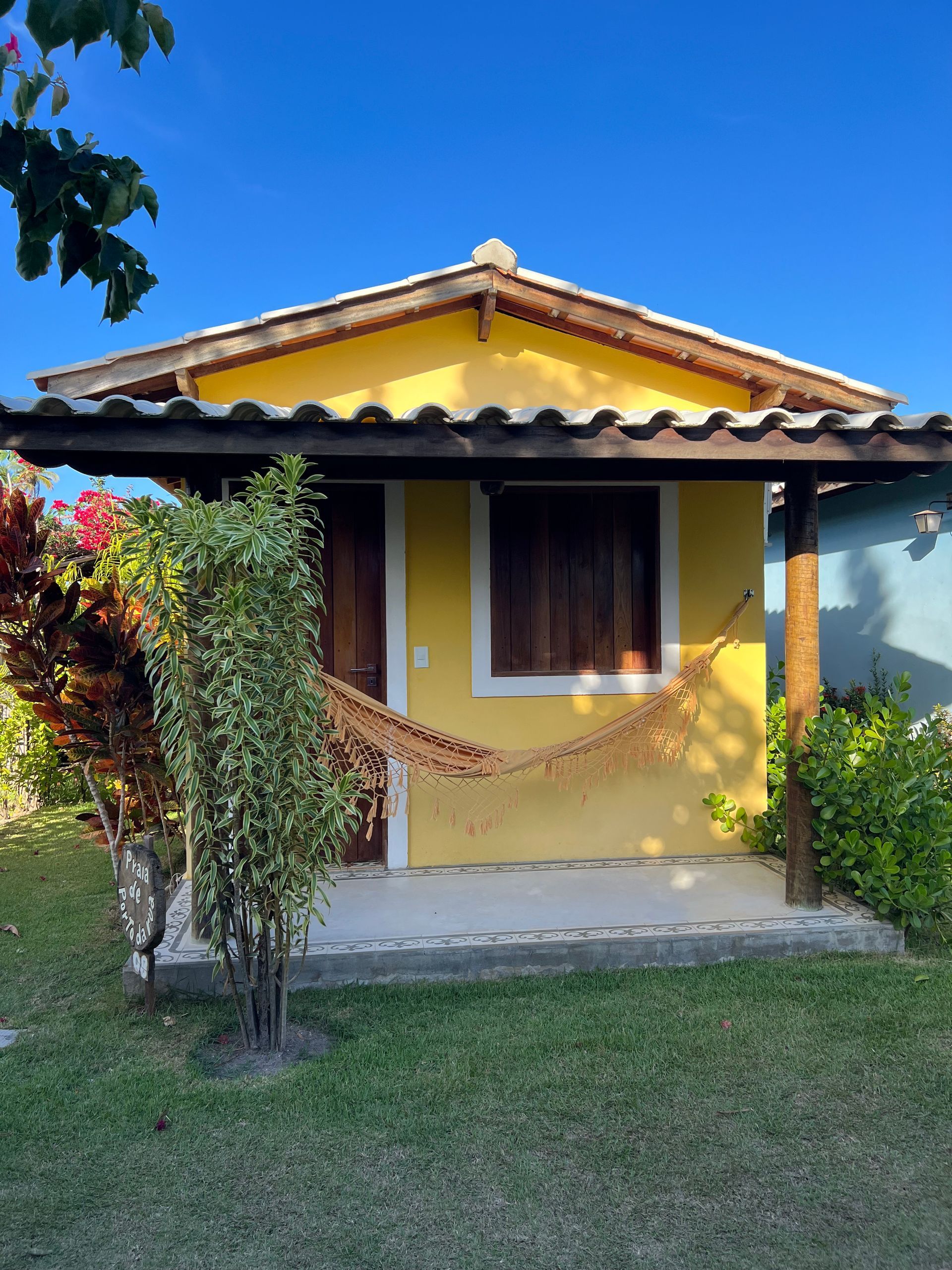Uma cabana amarela de um só andar, com telhado marrom e uma rede pendurada na varanda da frente, situada em uma área gramada e ensolarada.