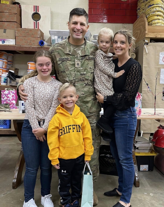 Family poses together; soldier in uniform with three children and woman. Smiling faces in a shop setting.