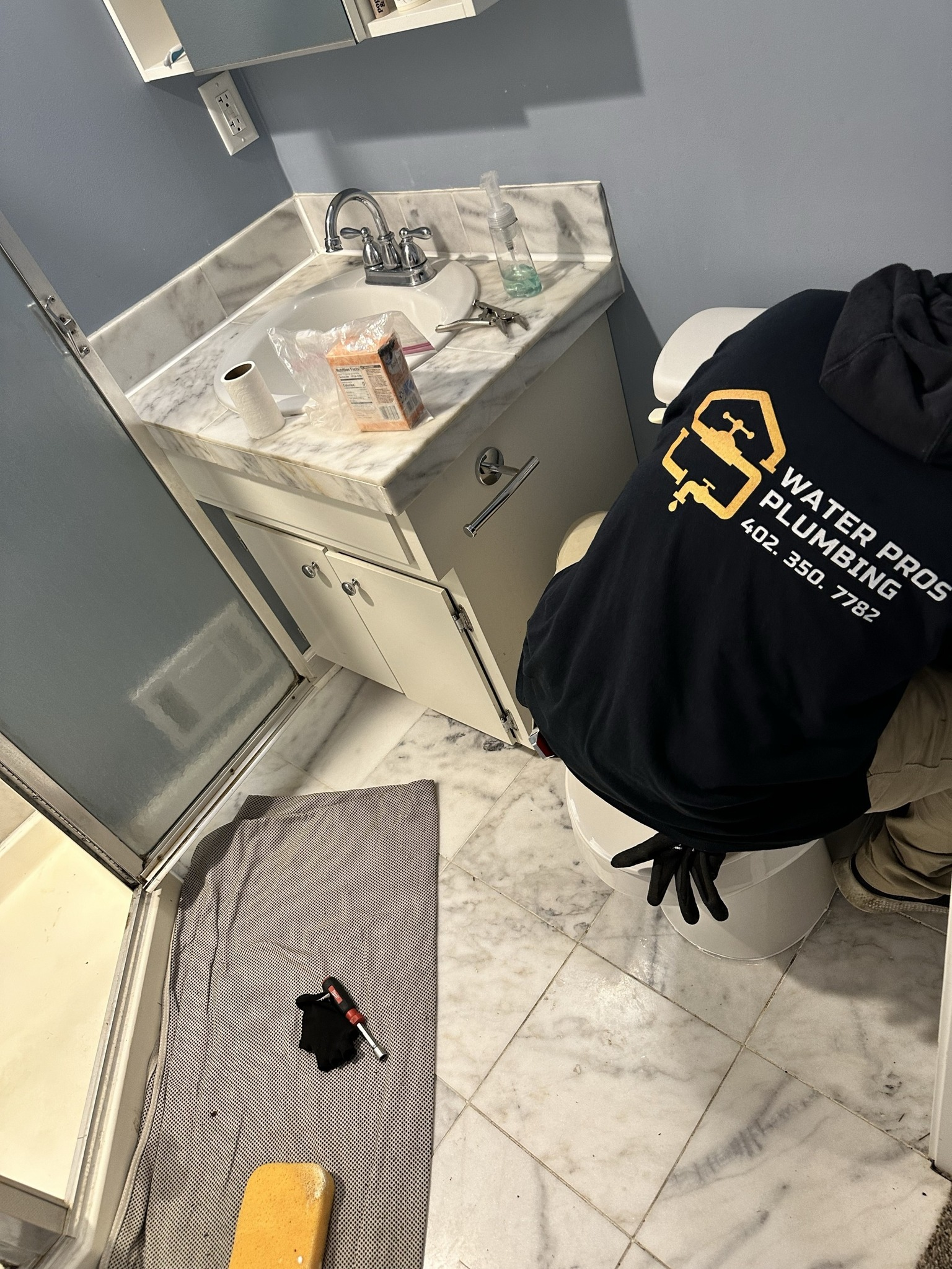 Plumber in bathroom fixing toilet. Black shirt with Water Pros Plumbing logo.