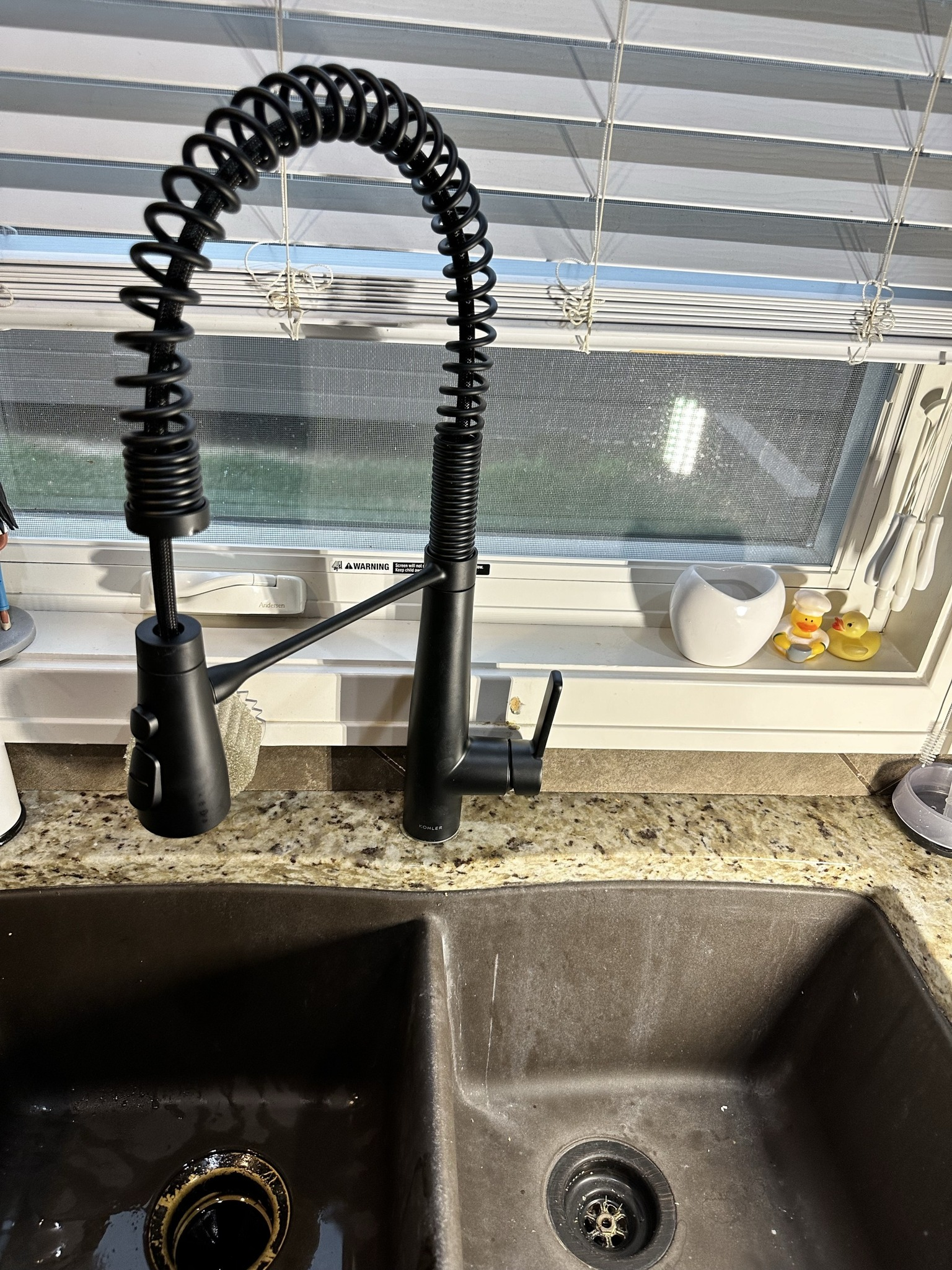 Black kitchen faucet with a coil, over a dark double sink. Blinds and a window behind it.
