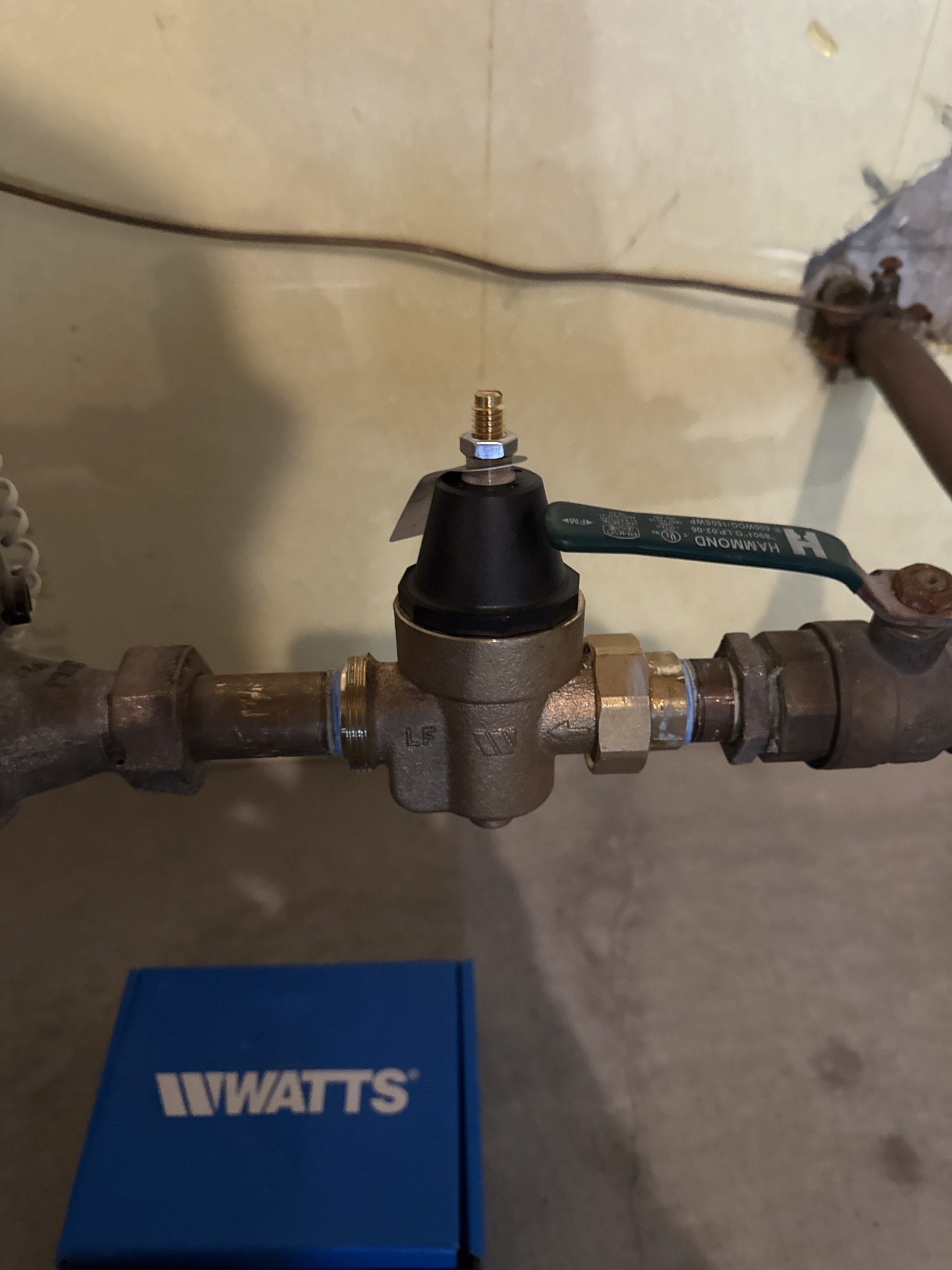 Brass water pressure regulator with black top and green handle, connected to pipes.