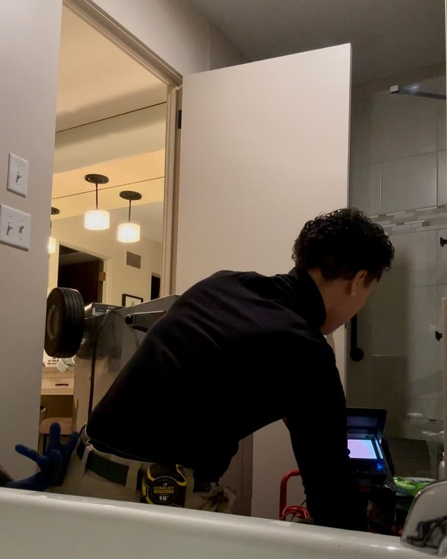 Person in black shirt using a device next to a bathtub; other room visible through doorway.