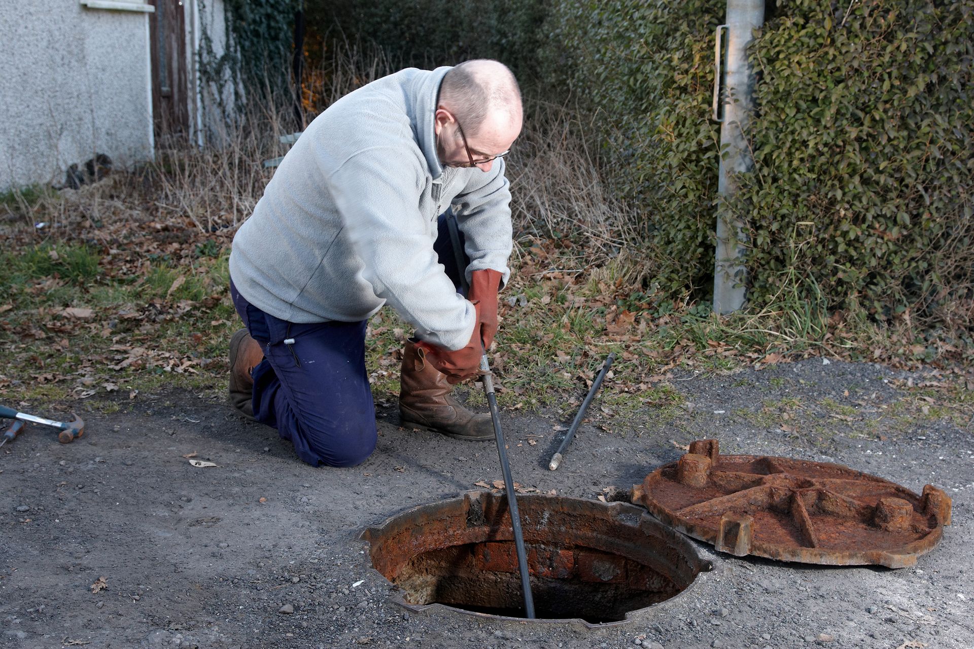 Worker inspecting and cleaning a manhole in an outdoor setting with tools on the ground.